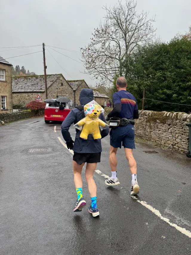 Sara Cox running with Pudsey on her back, Pudsey has a raincoat on