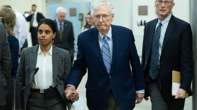 Mitch McConnell, in a dark suit, grips the arm of an aide as he walks through the Senate hallway