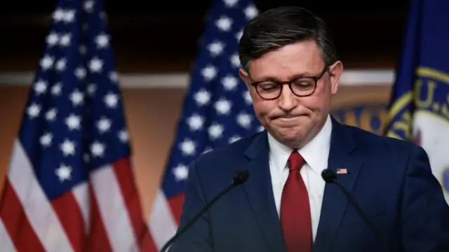 Speaker Mike Johnson, in a blue suit and red tie, looks down and frowns, against a backdrop of American flags