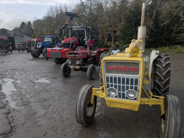 A line of tractors stood in a muddy field, at the front is a yellow tractor with a Pudsey teddy on top