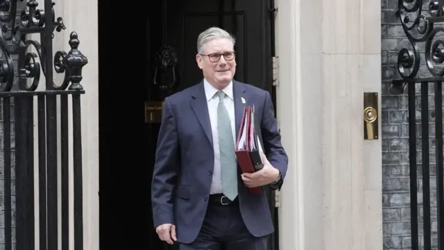 Starmer outside Downing Street, in a blue suit and green tie, he has grey hair and is holding two folders.