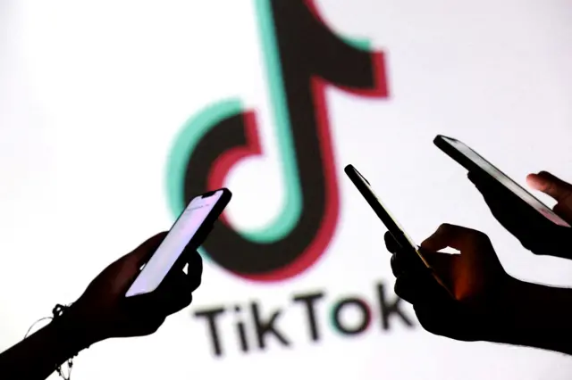 Three hands holding phones while a bright screen displays the TikTok logo (a black note with a t-shaped flag with red and blue contours) in the background