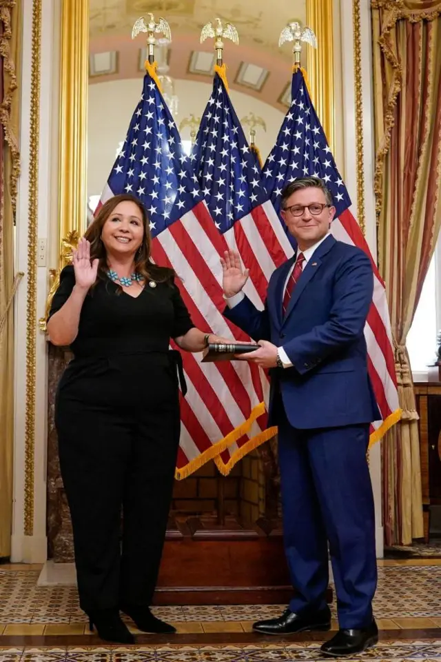 A woman with brown hair, dressed in black, and with her hand on a Bible is sworn into office by a man in a blue suit with glasses and a red tie