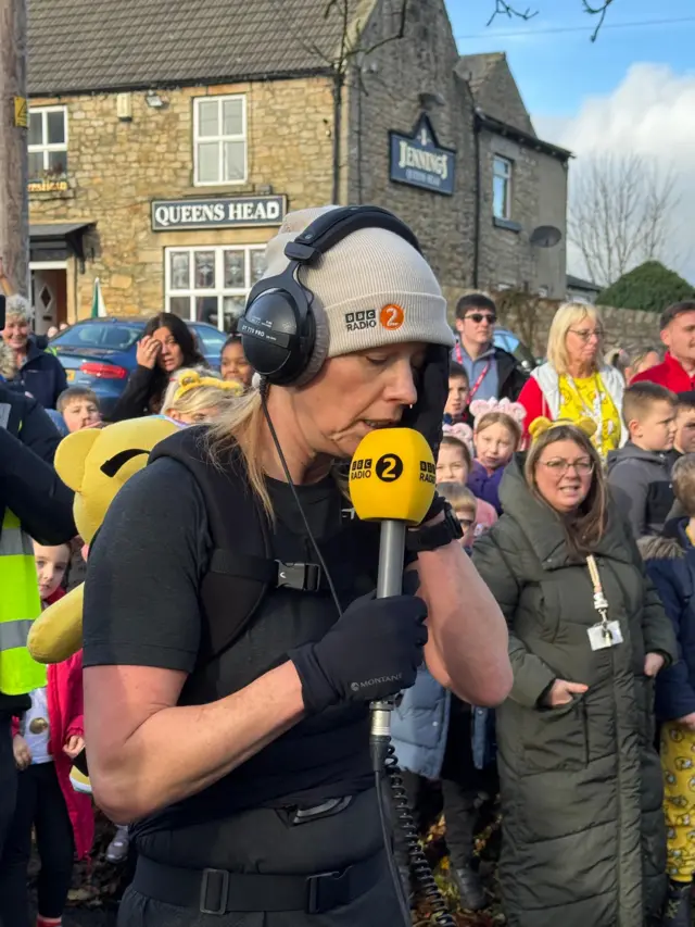 Sara Cox with her eyes closed and a hand to her face, looking tired. She has headphones on and is holding a microphone
