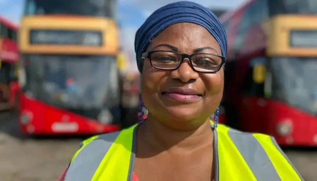 Woman with glasses and yellow high vis jacket smiling at the camera. Two New Routemaster double-decker buses are parked behind her