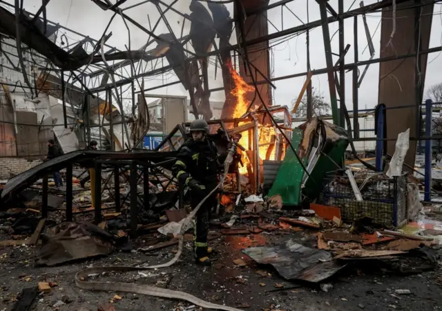 A firefighter holding a hose inside a damaged and burning warehouse