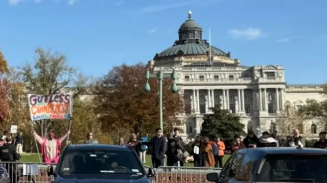 Outside the US Capitol, protesters are gathered. Some hold signs that mention Epstein and call for the release of more files