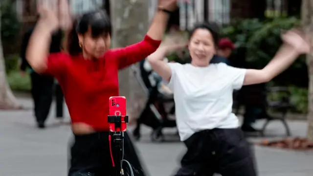 Two women in a park dance in front of a phone camera, propped on a tripod