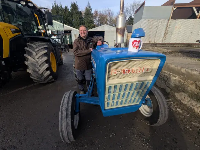A man posing next to a blue Ford tractor, with a mug sitting on top the tractor that says 'COX & A CUPPA'