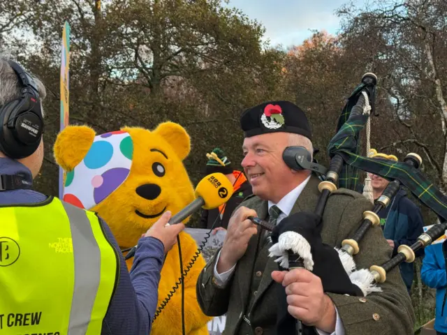 Pudsey stands with a man playing bagpipes