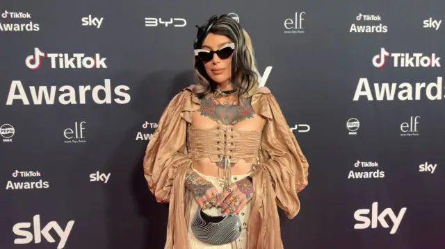 Sammi Jefcoat poses for pictures in a beige corset, white skirt and holding a black pouch