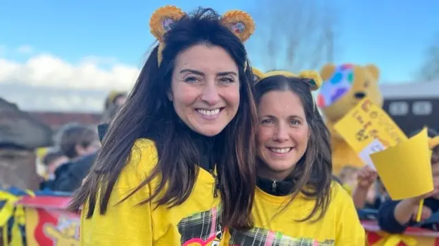 Two women with yellow t-shirts on and yellow headbands with ears on. There is a group of children in the background but they are blurred or obscured from view with yellow signs of encouragement.