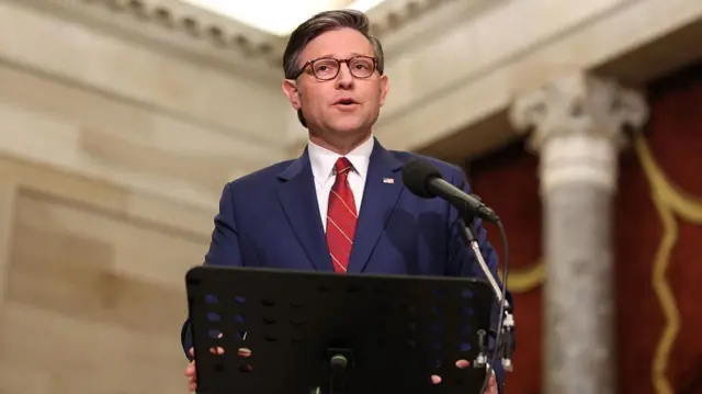 Mike Johnson speaks at a podium while wearing a navy suit and red tie