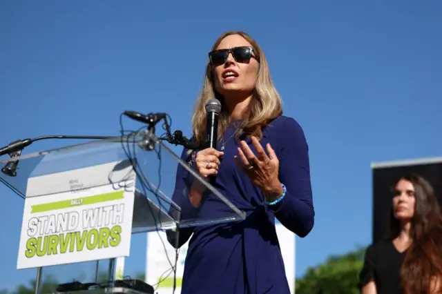 Annie Farmer speaking at a rally in front of a podium which says 'stand with survivors'