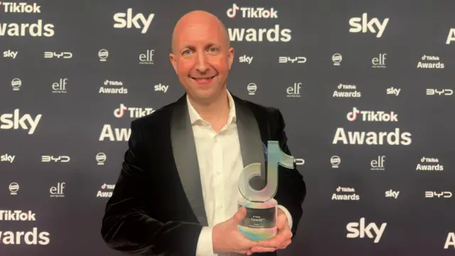 Phil holds his award up and smiles at the camera