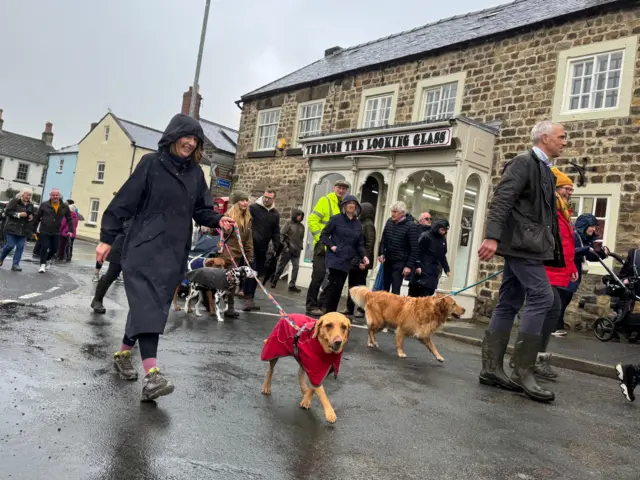 A woman smiling walking her dog in a raincoat, with some more dogs and owners behind