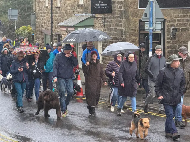 A crowd of people in raincoats and unbrellas, with a few dogs on leads