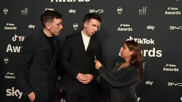 Matthew and Michael Gardiner (L and C) stand as BBC journalist Yasmin Rufo holds up a mic to them as she interviews them
