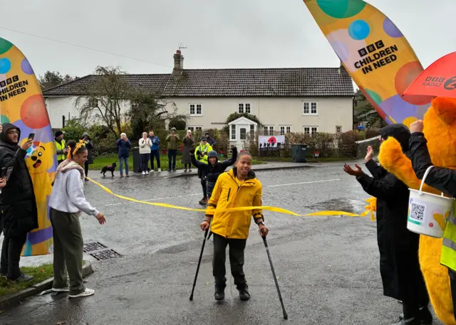 A young man crosses a finish line. He is holding crutches in each hand and has a yellow jacket on.