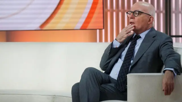Michael Wolff sat on a beige sofa on a TV set, with his hand placed by his mouth