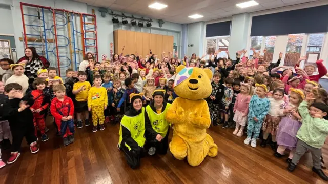Lindsey, Amy and Pudsey the big yellow mascot bear kneels down in front of a crowd of about 100 school children.