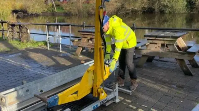 A man in a yellow coat and white helmet working on a yellow metal construction on a road with a metal barrier behind him