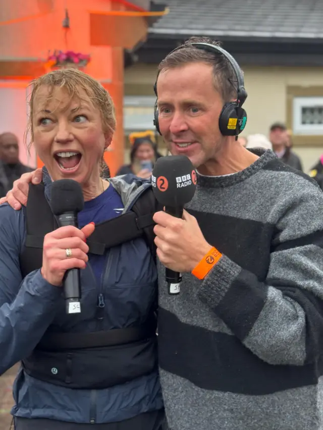 Sara Cox and Scott Mills hugging and holding microphones, looking happy