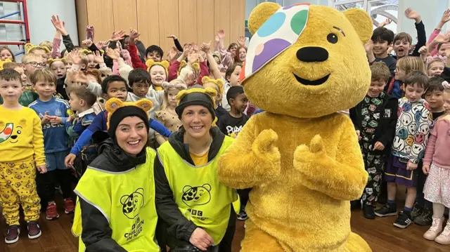 Lindsey, Amy and Pudsey the big yellow mascot bear kneels down in front of a crowd of about 100 school children.