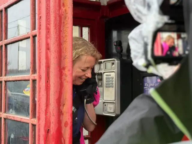 Sara smiles into a phone in a phone box