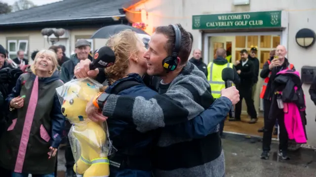 Sara Cox and Scott Mills hugging outside Calverley Golf Club, with people behind them smiling