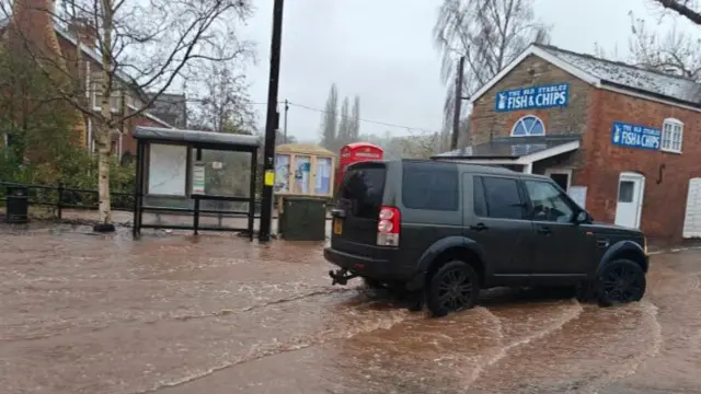 A black four wheel drive vehicle driving through muddy water with a fish and chip shop in the background
