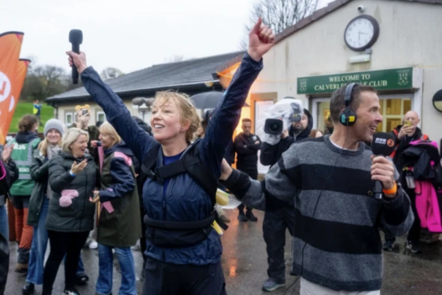 A woman with blond hair in a plait or ponytail holds her hands up in celebration. She is wearing a navy rain coat and a rucksack.