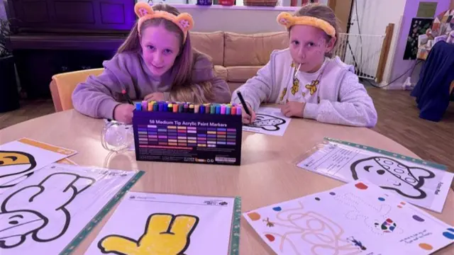 Two young girls with yellow bear ear headbands on are colouring in pictures of Pudsey the bear.