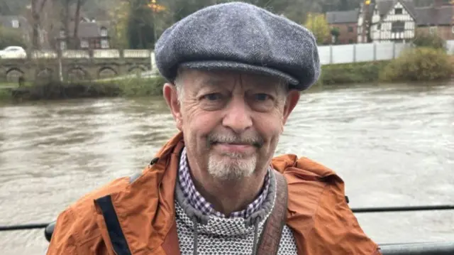 A man with a grey cap and an orange coat standing in front of a river