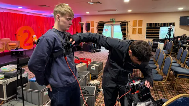 A person wearing a camera on a vest and a second man testing some wires