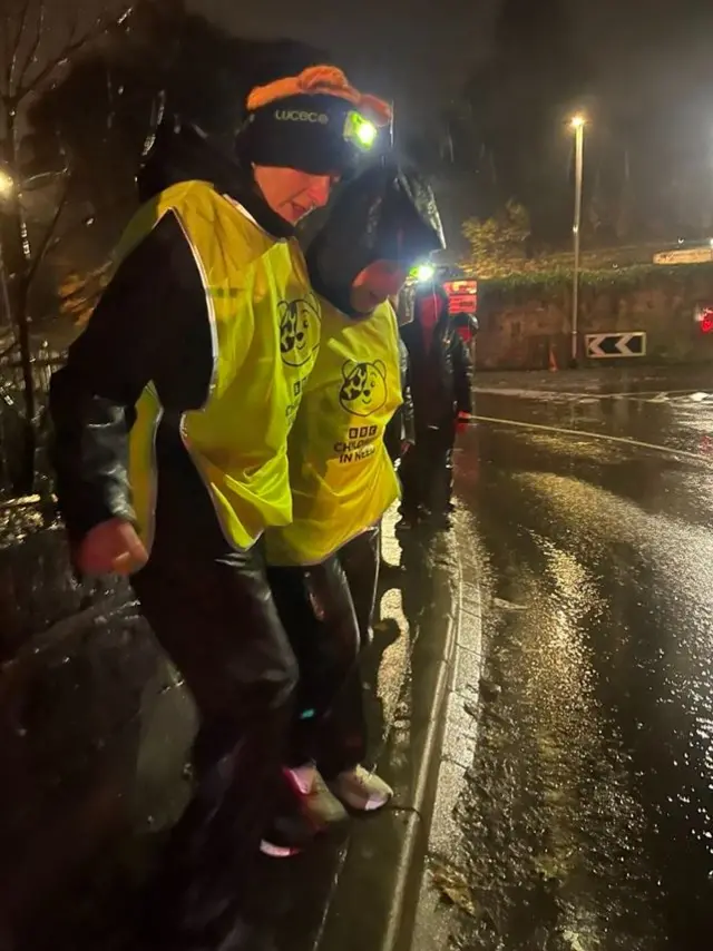 Two women wearing waterproofs, high-vis jackets and head torches are walking side by side on a narrow pavement. People can be seen behind them. It is a dark and rainy evening.