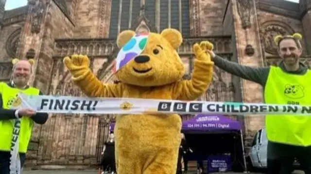 Pudsey, a yellow bear mascot with a spotted handkerchief over one eye, crosses a finish line which reads "BBC Children in Need".