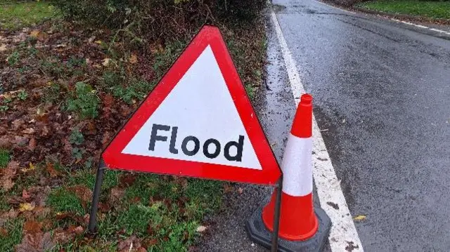 A red and white warning triangle with the word flood on it and a road behind it