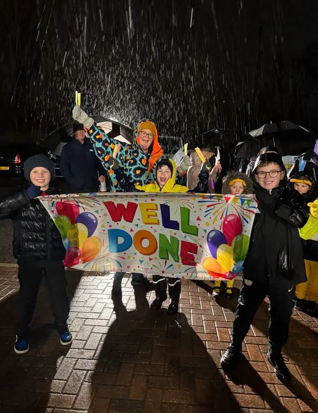 A woman and four children hold up a banner that says Well Done. They are dressed in warm coats and are outside in the dark and it is raining.