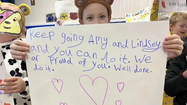 A girl holds up a hand-drawn poster. She looks at the camera but most of her face is covered. It saus "Keep going Amy and Lindsey and you can do it. We are proud of you. Well done. Do it". Pink heats are drawn underneath it.