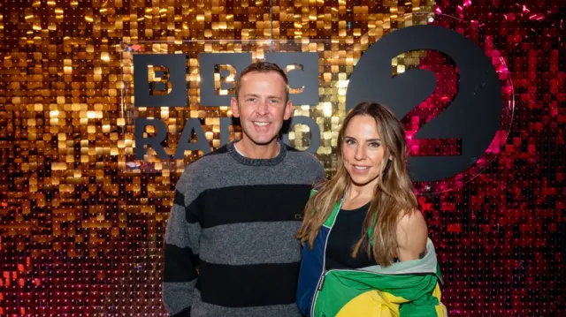 Scott Mills and Mel C smiling and posing in front of a sparkly wall with the BBC Radio 2 logo