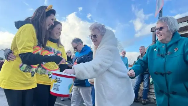 Amy and Lindsey, both with mid-length brown hair and wearing yellow t-shirts and yellow bear ears, accept donations in a bucked from two elderly women who are smiling.