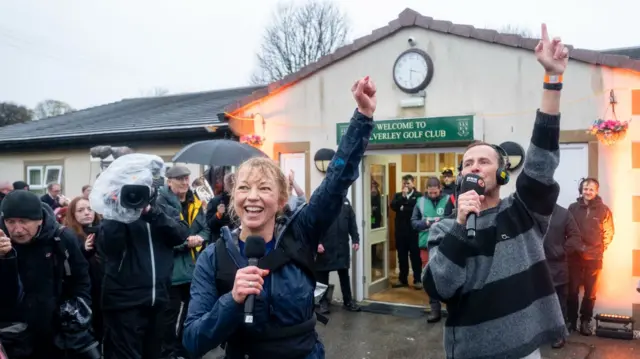 Sara Cox smiling with her arm in the air stood next to Scott Mills holding a microphone with his arm in the air, stood outside Caverley Golf Club