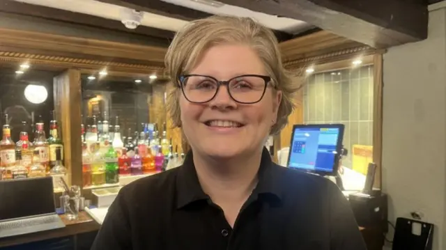 A woman with blonde hair and dark rimmed glasses in a pub