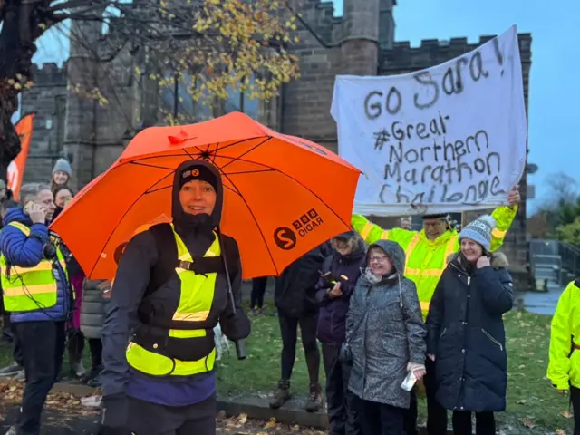 Sara Cox holding a bright orange umbrella  with a crowd.