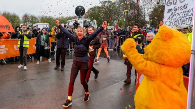 Sara Cox smiling with her arms in the air at the finish line of her Children in Need challenge, a crowd is around her cheering and there's confetti in the air