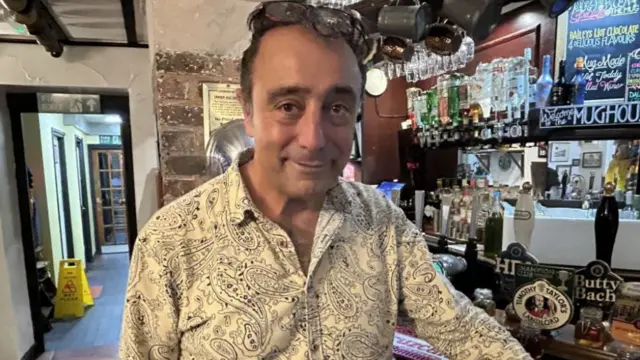 A man with dark hair and a white patterned shirt standing at a bar with a number of pumps running along it