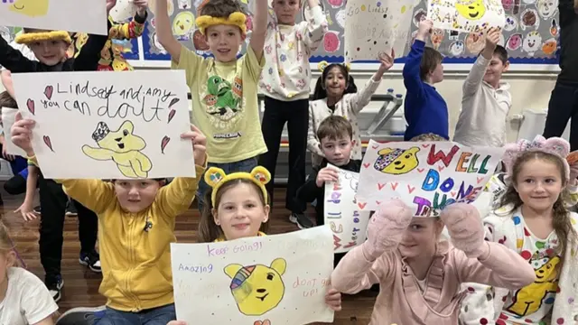 Lots of children hold up posters with "you can do it" and "well done" written on them. Lots of them have drawings on of the yellow Pudsey bear. Most of the children are wearing headbands with bear ears on the,m.