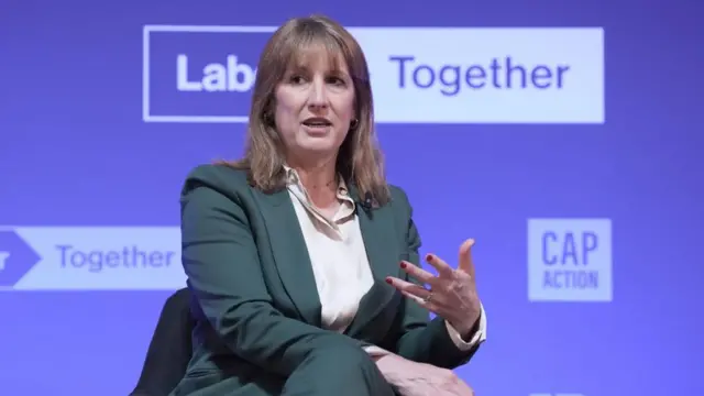 Rachel Reeves on stage wearing a dark green suit and white shirt, gesturing with one hand, in front of a blue backdrop