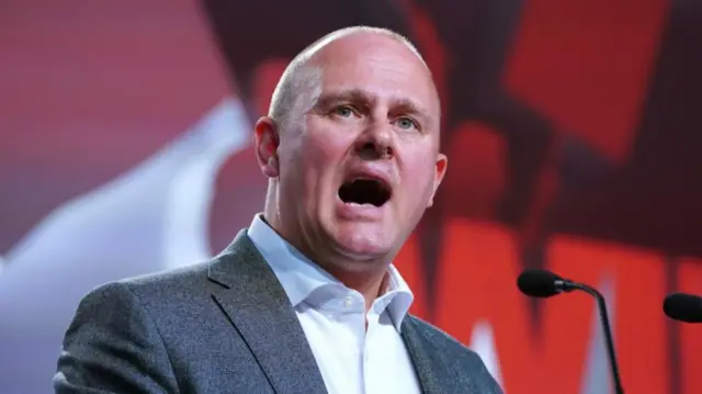 A man in a grey jacket with a white shirt standing at a microphone